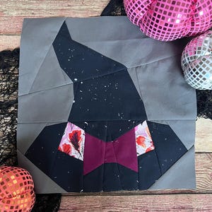 May include: A fabric block featuring a geometric design of a witch's hat. The hat is black with white speckles and a burgundy bow. The brim has floral accents. The background is gray. Decorative pink and silver pumpkins are in the background.
