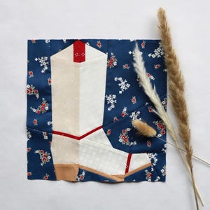 Puede incluir: Una obra de arte textil que representa una bota de vaquero blanca con detalles rojos, sobre un fondo azul con estampado floral. La composición cuadrada incluye hierbas secas. El diseño de la bota es de estilo patchwork.