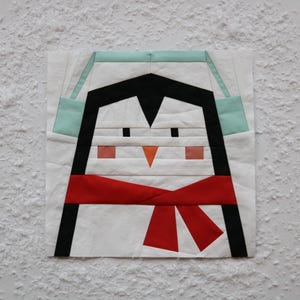 Puede incluir: Un pingüino blanco con una bufanda roja y un gorro azul claro. El pingüino tiene ojos negros, un pico naranja y mejillas rosadas.