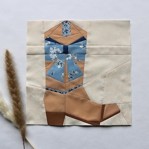 Op de afbeelding: Een stoffen blok met een patchwork ontwerp van een cowboylaars. De laars is voornamelijk bruin met blauwe bloemenaccenten en een donkerblauwe strik. De achtergrond is lichtbeige met een subtiel bloemenpatroon.
