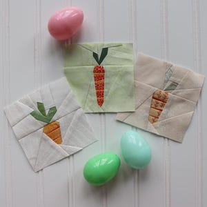 Puede incluir: Tres cuadrados de tela con forma de zanahoria con tapas verdes y cuerpos naranjas. Los cuadrados están sobre una superficie blanca con tres huevos de Pascua de plástico, dos verdes y uno rosa.