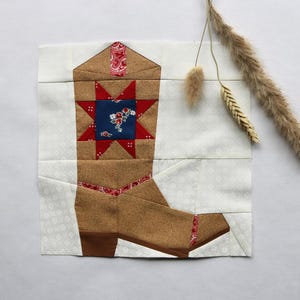 Könnte beinhalten: Ein Patchwork-Design einer Cowboy-Stiefel. Der Stiefel ist hauptsächlich braun mit roten und blauen Akzenten, einschließlich eines Sternmotivs. Der Hintergrund ist weiß mit einem zarten Blumenmuster. Die quadratische Form eignet sich für einen Quiltblock oder eine Wanddekoration.