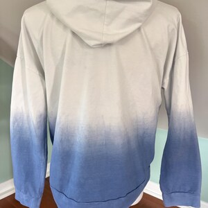 May include: A white and blue ombre hoodie with a drawstring hood. The long-sleeved sweatshirt transitions from white at the top to blue at the bottom. The hem and cuffs are blue.