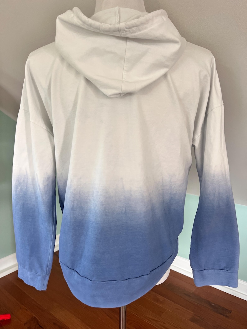May include: A white and blue ombre hoodie with a drawstring hood. The long-sleeved sweatshirt transitions from white at the top to blue at the bottom. The hem and cuffs are blue.