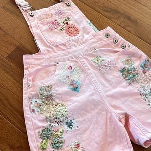 May include: Pink denim overalls with patchwork details. The overalls feature floral and lace patches, a front pocket, and adjustable straps. The patches include various floral patterns and textures, adding a unique, handcrafted look.