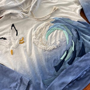 May include: White hooded sweatshirt with a blue ombre effect. The design features a seagull and a wave made of various blue fabrics. The sleeves are solid blue. The hoodie has a drawstring and a small tag.