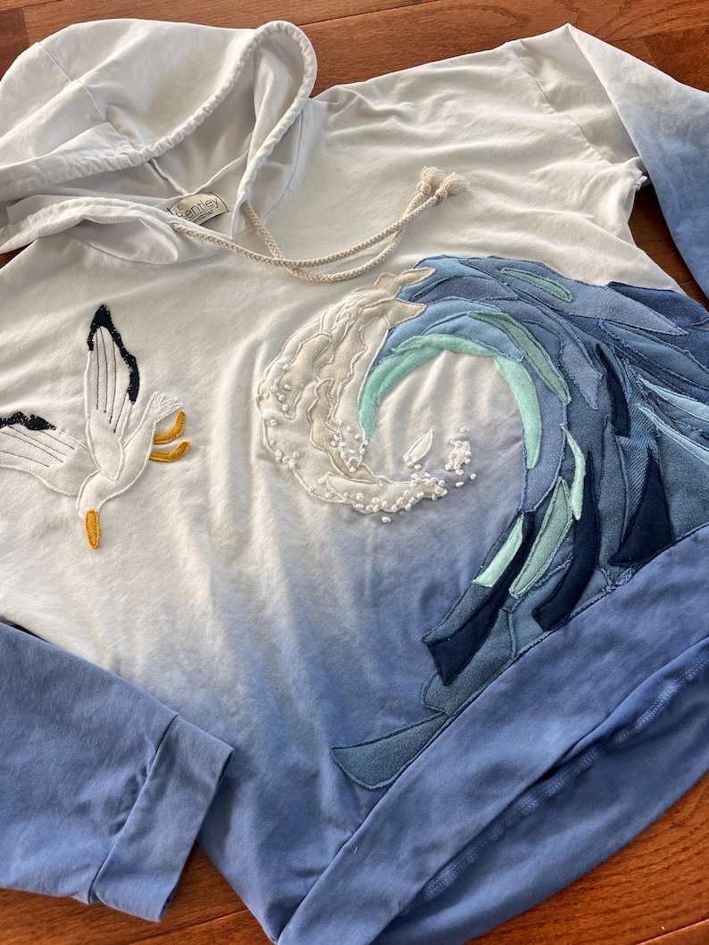 May include: White hooded sweatshirt with a blue ombre effect. The design features a seagull and a wave made of various blue fabrics. The sleeves are solid blue. The hoodie has a drawstring and a small tag.