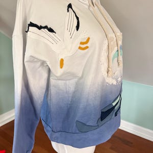 May include: White hooded sweatshirt with a gradient blue design. The sweatshirt features an embroidered seagull and other nautical-themed appliqu&eacute;s. The drawstring is a cream color. The sleeves are long and the bottom hem is a solid blue.