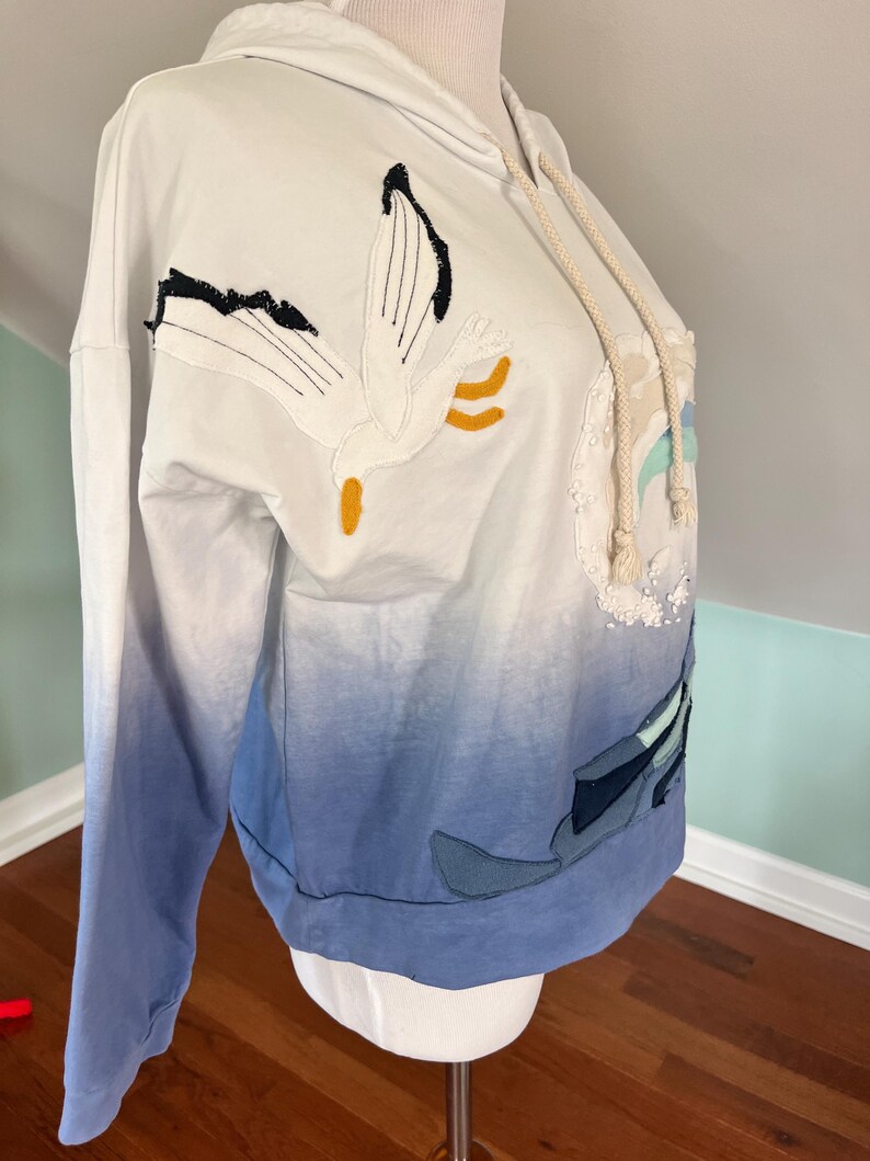 May include: White hooded sweatshirt with a gradient blue design. The sweatshirt features an embroidered seagull and other nautical-themed appliqu&eacute;s. The drawstring is a cream color. The sleeves are long and the bottom hem is a solid blue.