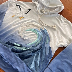 May include: White hoodie with a blue ombre design. Features a seagull applique and a wave design made of various shades of blue fabric. The hoodie has a drawstring and long sleeves.