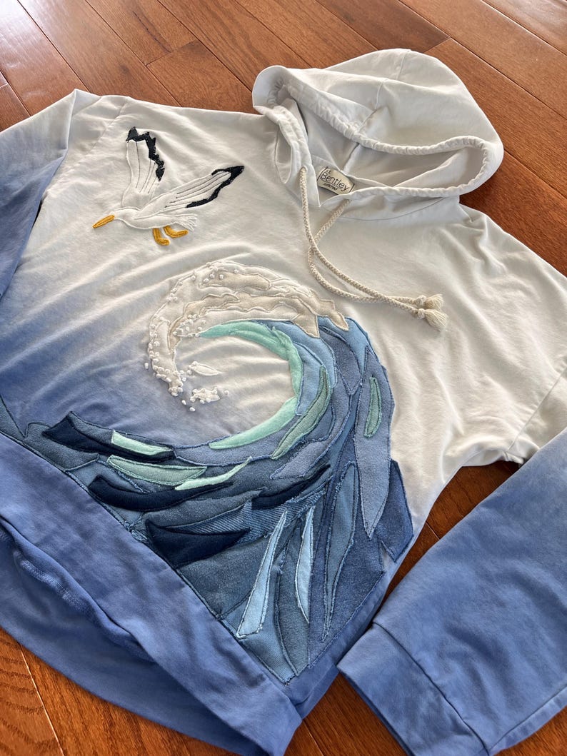 May include: White hoodie with a blue ombre design. Features a seagull applique and a wave design made of various shades of blue fabric. The hoodie has a drawstring and long sleeves.
