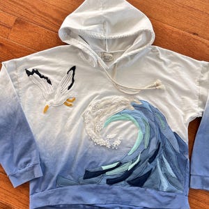 May include: White hooded sweatshirt with a blue ombre design. Features an embroidered seagull and a wave design with blue and white fabric patches. The brand name "Bentley" is visible on the label.