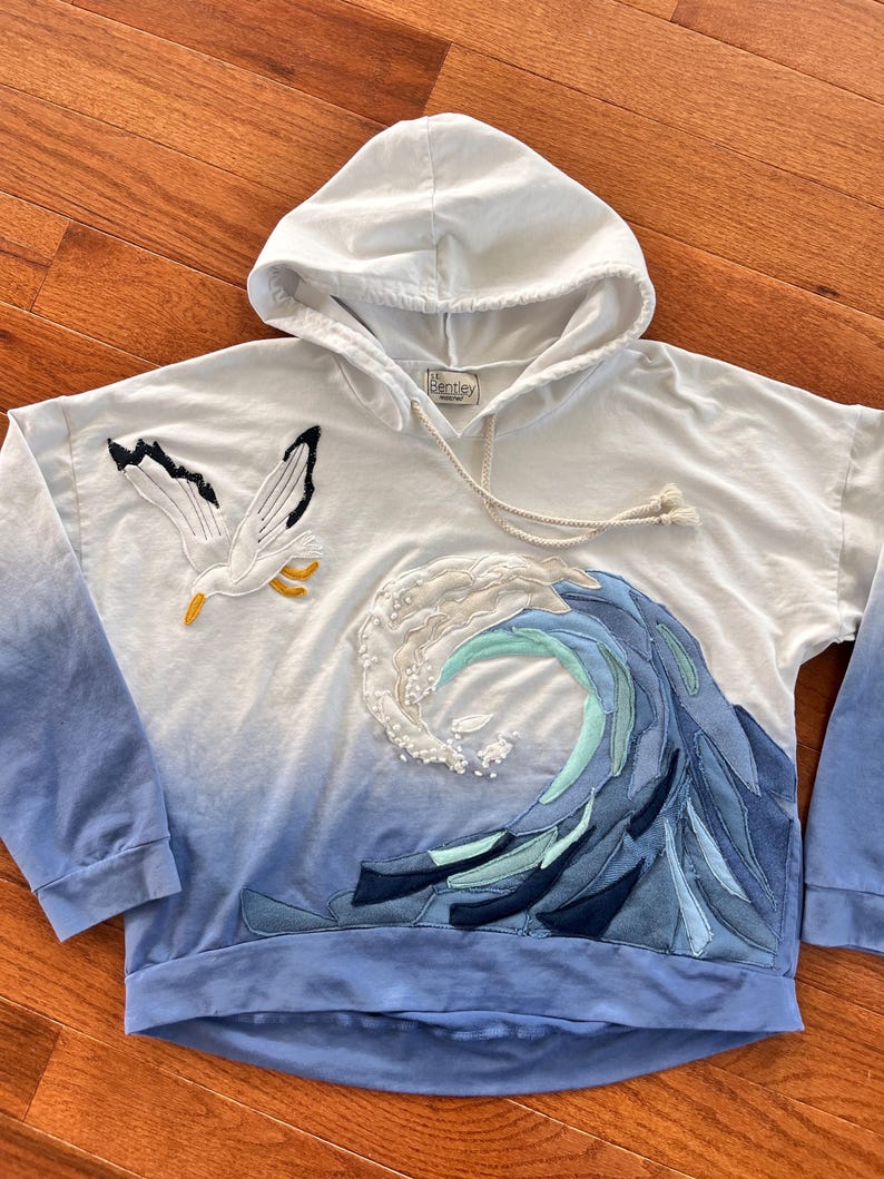 May include: White hooded sweatshirt with a blue ombre design. Features an embroidered seagull and a wave design with blue and white fabric patches. The brand name "Bentley" is visible on the label.