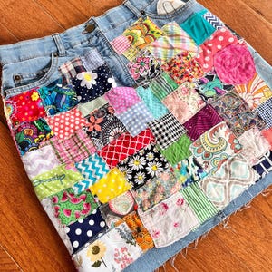 May include: Light blue denim skirt with a patchwork design. The patchwork is made of various colorful fabric squares with floral, polka dot, and geometric patterns. The word "Gossip" is visible on the skirt.