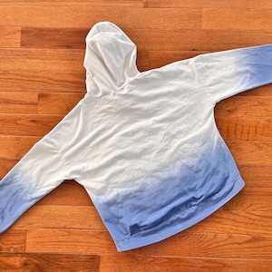 May include: A white and blue ombre hoodie with a hood and long sleeves. The hoodie transitions from white at the top to a light blue at the bottom. The garment is made of a soft material and is laid flat on a wooden surface.