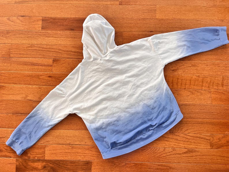May include: A white and blue ombre hoodie with a hood and long sleeves. The hoodie transitions from white at the top to a light blue at the bottom. The garment is made of a soft material and is laid flat on a wooden surface.