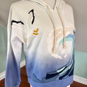 May include: White hoodie with a blue ombre effect on the sleeves and lower body. The hoodie features an embroidered design of a wave and a flower. The drawstring is beige.