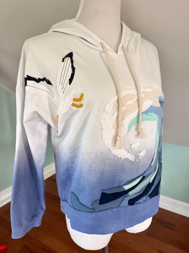 May include: White hoodie with a blue ombre effect on the sleeves and lower body. The hoodie features an embroidered design of a wave and a flower. The drawstring is beige.