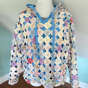 May include: A unique hoodie featuring a patchwork design of colorful fabric circles. The hoodie has a blue lining, a drawstring hood, and long sleeves. The patchwork includes various colors like red, pink, purple, and cream.