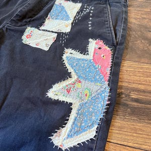 May include: Dark blue denim pants with patchwork details. The patchwork is made of various floral fabrics in shades of blue, pink, and white. The pants have a single front pocket.
