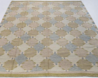 May include: A rectangular area rug featuring a repeating diamond pattern in shades of beige, light blue, and cream. The design consists of interlocking geometric shapes with small dark blue accents at the corners, finished with a solid olive green border.