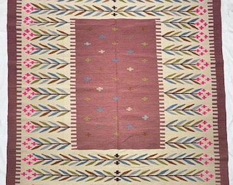 May include: A rectangular rug with a central maroon field and a cream-colored border. The border features a repeating pattern of blue, green, and brown leaves with pink floral accents. The rug's edges are trimmed with a fringe.