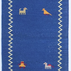 Può includere: Un tappeto blu rettangolare con bordo a zig-zag color crema e frange. Il tappeto presenta immagini ricamate di uccelli e animali in giallo, rosso e crema. Un elemento decorativo per la casa.