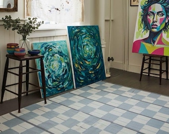 May include: A blue and white chequered rug in a room with artwork. Two abstract paintings lean against the wall, and a portrait of a woman stands on an easel. A small wooden table holds decorative bowls.
