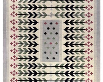 May include: A cream-colored rug with a geometric pattern of black arrows, green leaves, and red crosses. The rug has a light gray border with a green stripe.