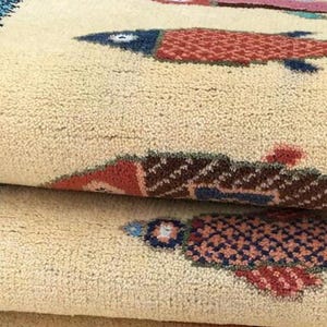 May include: Close-up of a beige rug with a repeating pattern of stylized fish. The fish are in shades of red, blue, and brown, with a textured, woven appearance. The rug is folded, showing multiple layers.