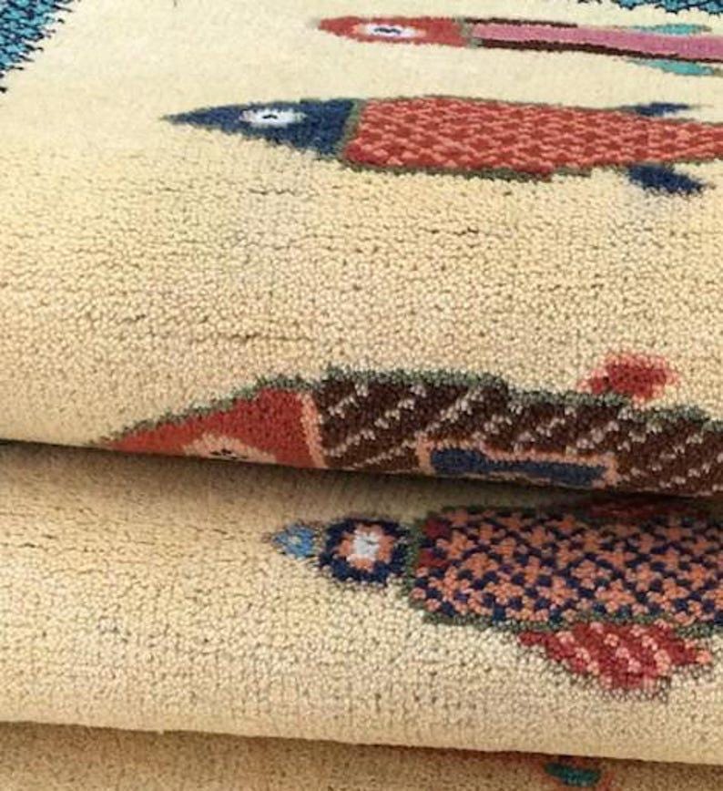 May include: Close-up of a beige rug with a repeating pattern of stylized fish. The fish are in shades of red, blue, and brown, with a textured, woven appearance. The rug is folded, showing multiple layers.