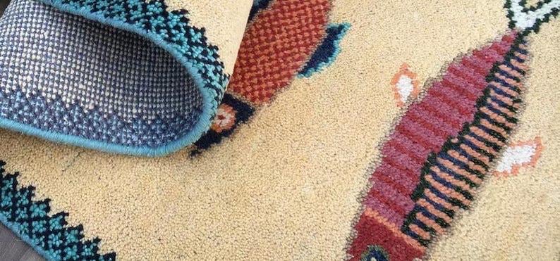 May include: Beige rug with a fish design. The fish is primarily red with black and orange stripes. The rug's border is teal and navy blue with a geometric pattern. The underside of the rug is visible, showing a navy blue mesh.