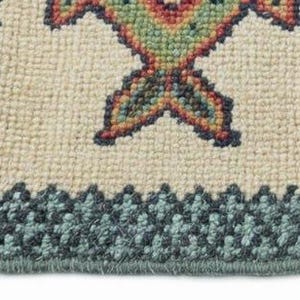 May include: Close-up of a beige rug with a geometric pattern in shades of green, red, and orange. The rug has a dark teal border and fringe on one side. The rug's design features a repeating motif.