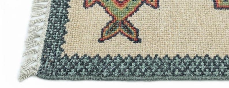 May include: Close-up of a beige rug with a geometric pattern in shades of green, red, and orange. The rug has a dark teal border and fringe on one side. The rug's design features a repeating motif.