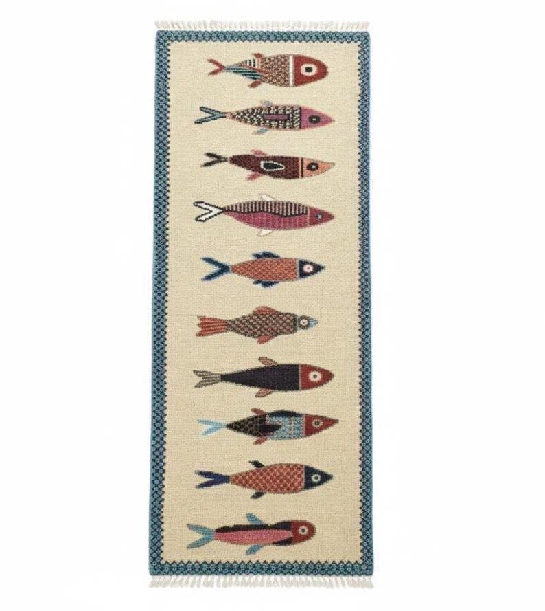 May include: A rectangular rug with a cream-colored background and a border of blue geometric patterns. The rug features a repeating pattern of stylized fish in various colors, including red, pink, and black. The rug has a fringed edge.