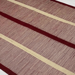 Può includere: Un tappeto tessuto con un motivo a strisce in tonalità bordeaux e beige. Il tappeto ha una superficie testurizzata e un bordo con frange.