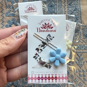 May include: A set of three hair clips with floral patterned tape and a blue flower charm. The packaging includes the text "Tatreez With Danduna" and an Instagram handle.