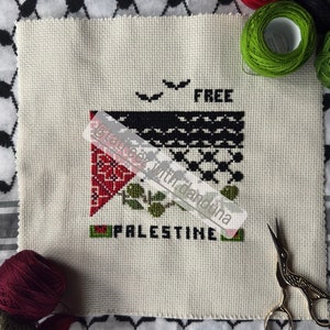 Może przedstawiać: Haftowane krzyżykiem palestyńskie flagi z napisem "FREE PALESTINE" i dwoma ptakami lecącymi nad nią. Haftowane na białym materiale i otoczone czerwoną, zieloną i brązową nicią.