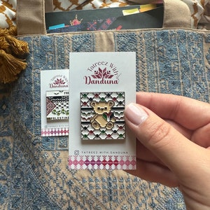 May include: A white card with the text "Tatrezz with Danduna" in maroon lettering. The card features a small, pixelated, beige bear with a red flower in its paw. The bear is surrounded by a colourful, pixelated design. The card is on a blue and beige patterned fabric.