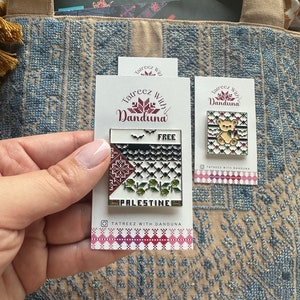 Peut inclure: Deux épingles en émail avec un fond blanc et des motifs de broderie colorés. La première épingle représente un drapeau palestinien avec le mot "Palestine" et le texte "Tatreez With Danduna" en dessous. La deuxième épingle représente un ours de dessin animé portant un keffiyeh palestinien et le texte "Tatreez With Danduna" en dessous.