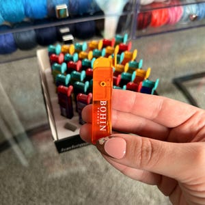 May include: An orange Bohin France needle threader held in a hand, with the brand name visible. The threader is rectangular with a small loop at the top. A display of colorful needle threaders is in the background, along with spools of yarn.