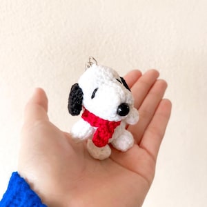Puede incluir: Llavero de perro blanco tejido a crochet con ojos negros y una bufanda roja.