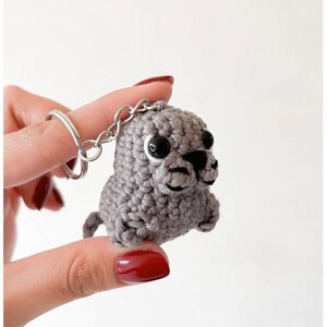May include: A small, gray crocheted seal keychain featuring a textured yarn body, two round black plastic eyes, and a stitched black nose and mouth detail. The figure is attached to a silver-toned metal split ring for keys or accessories.