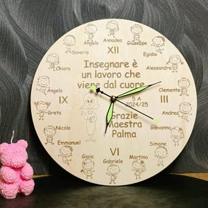 Puede incluir: Un reloj redondo de madera clara. La esfera del reloj presenta ilustraciones grabadas de niños y una maestra. Las manecillas del reloj son verdes. El texto en el reloj dice "Insegnare è un lavoro che viene dal cuore" y "Grazie Maestra Palma."