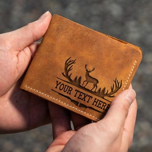 May include: A brown leather wallet with a deer silhouette and antlers engraved on the front. The text "YOUR TEXT HERE" is also engraved on the wallet.
