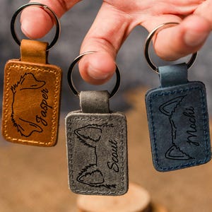 May include: Three leather keyrings with metal rings. Each keyring is a different colour: brown, grey, and blue. The keyrings are engraved with a fox silhouette and a name: Jasper, Scout, and Mochi.