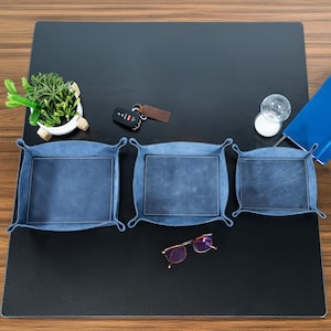 May include: Three blue leather catchall trays of varying sizes are arranged on a black surface. The trays have a square shape with a darker blue leather center. A small plant, keys, glasses, and a notebook are also on the surface.