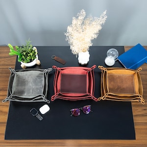 May include: Three leather valet trays in gray, red, and tan, each with a contrasting center panel. The trays are square with decorative corner stitching. The scene includes a small plant, a vase of dried flowers, and other desk accessories.