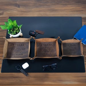 May include: Three leather trays with personalized text, each representing a different anniversary year. The trays are brown with dark brown leather inserts. The text includes names, dates, and milestones. A car key, glasses, and a small plant are also visible.