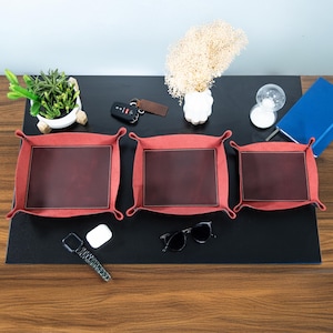 May include: Three red and brown leather catchall trays of varying sizes are arranged on a black surface. The trays have a rectangular shape with a brown leather center and red fabric border. Other items on the surface include sunglasses, keys, and a small plant.